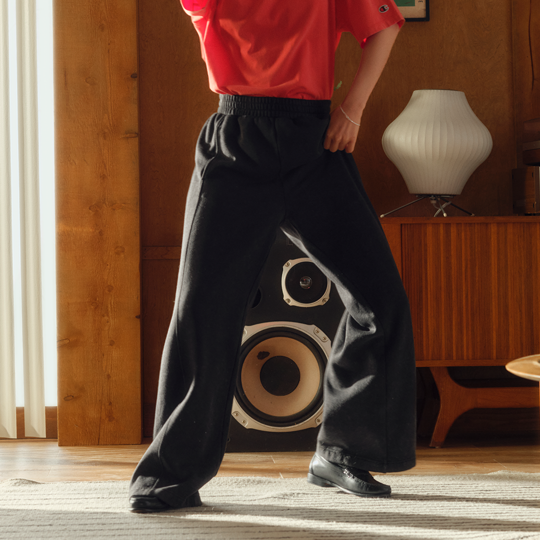 Person wearing black pants with a speaker design, standing in a room with wooden paneling and a lamp.