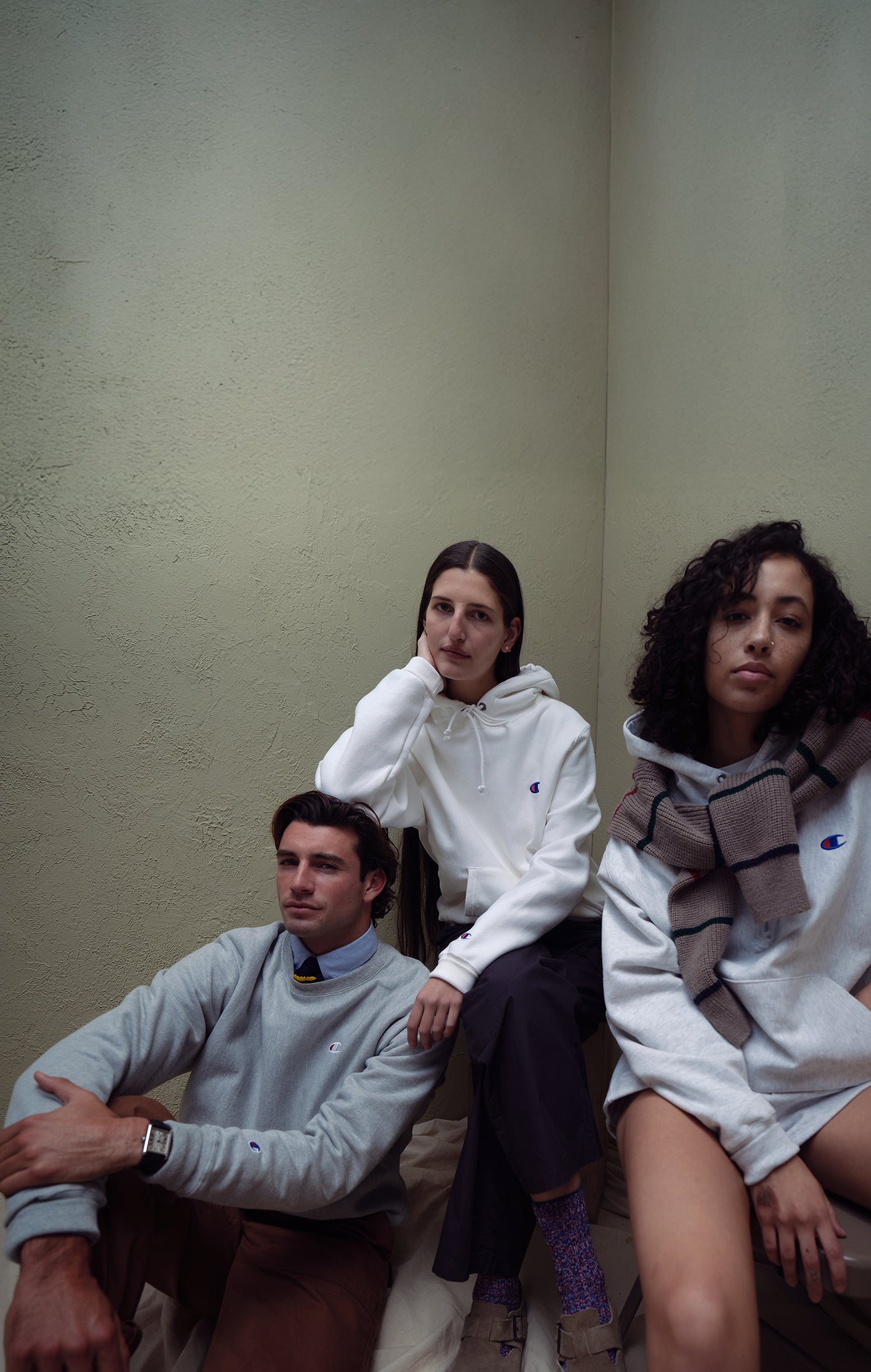 Three people sitting against a plain wall wearing champion sweatshirts and hoodies
