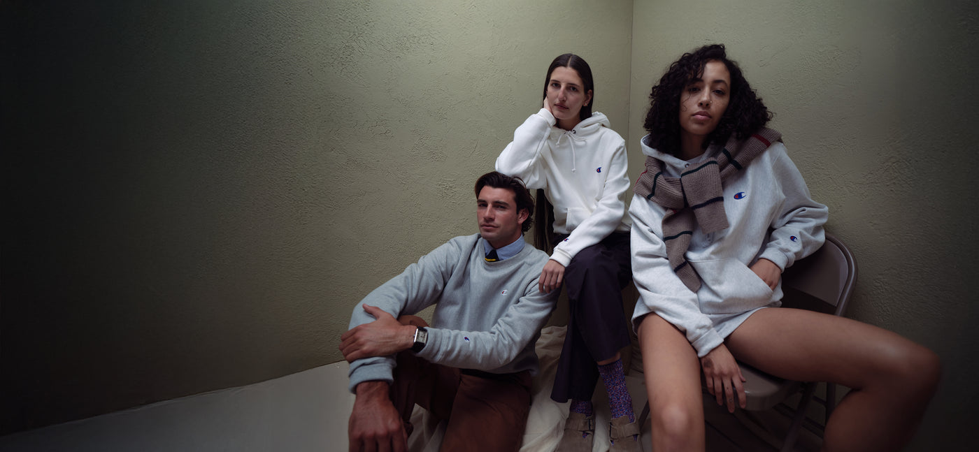 Three people sitting against a plain wall wearing champion sweatshirts and hoodies