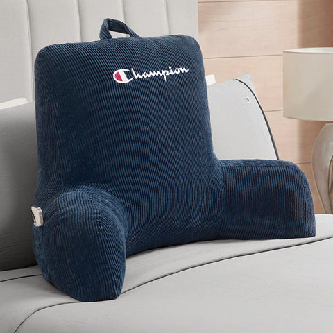 A Champion Corduroy Backrest Pillow in navy blue, featuring armrests, a built-in side pocket, and the Champion logo, is displayed on a bed with gray bedding and a lamp in the background.