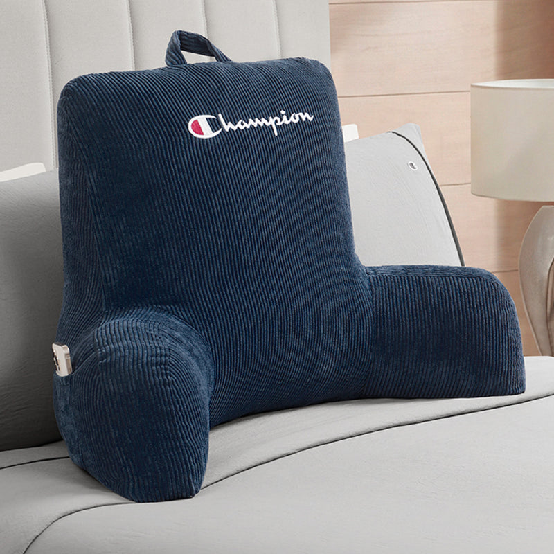A Champion Corduroy Backrest Pillow in navy blue, featuring armrests, a built-in side pocket, and the Champion logo, is displayed on a bed with gray bedding and a lamp in the background.