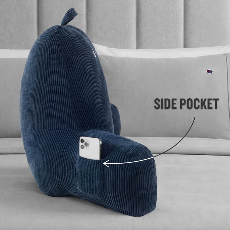 Champion Corduroy Backrest Pillow in dark blue, featuring a built-in side pocket that holds a phone, sits on a light-colored bed. An arrow and "SIDE POCKET" text highlight the functional pocket. Brand: Champion.