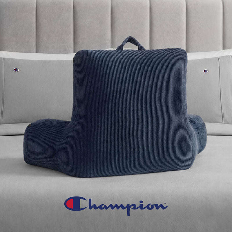 A Champion Corduroy Backrest Pillow in navy blue, featuring arms and a built-in side pocket, rests on a grey bed with Champion logo bedding. The Champion logo is also visible on the bedspread in front.