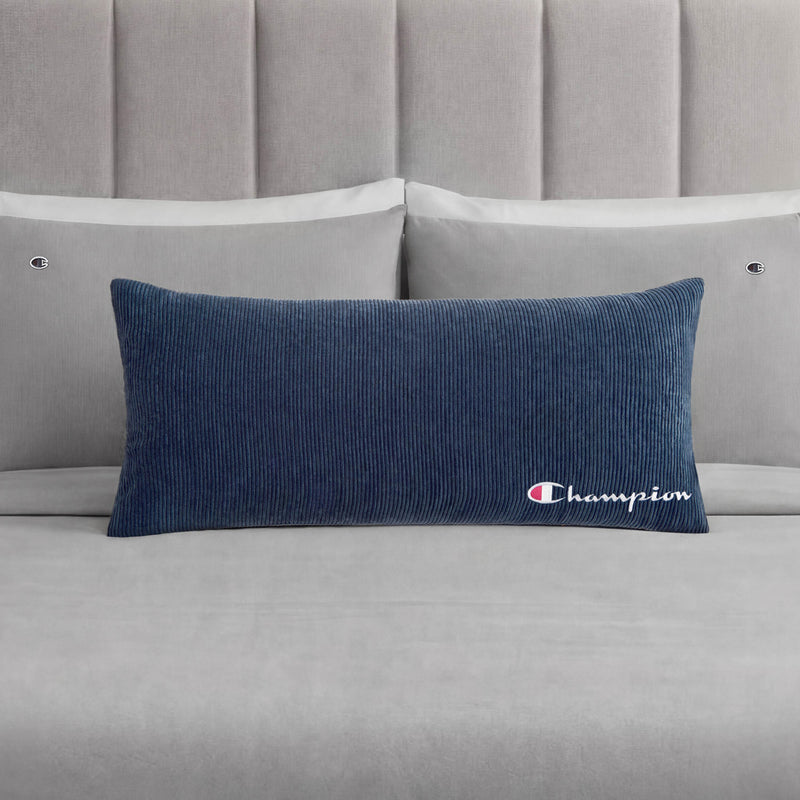 A Champion Corduroy Body Pillow in blue, featuring an embroidered logo, rests on a neatly made bed with light grey sheets, two grey pillows, and a padded grey headboard—creating the perfect lounge space to experience Champion comfort.