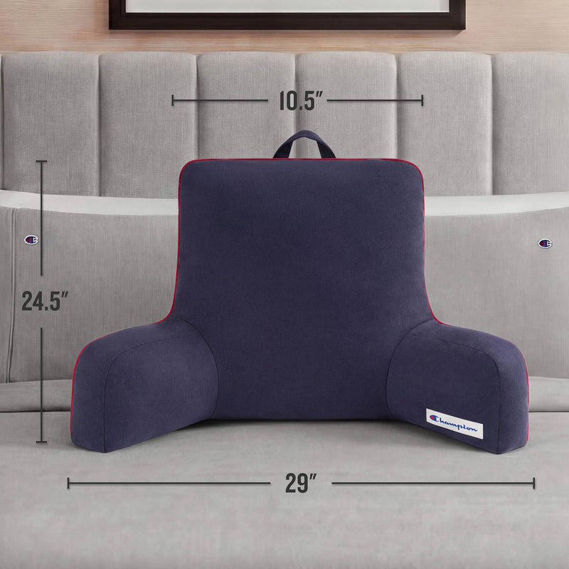 A Champion Faux Wool Backrest Pillow in dark blue with armrests is placed on the bed. Measuring 29" wide, 24.5" tall, and 10.5" deep, it features a convenient handle on top. The bed also has a light headboard and two Champion-branded pillows. This cozy backrest pillow from Champion offers extra comfort and support for reading or relaxing in bed.