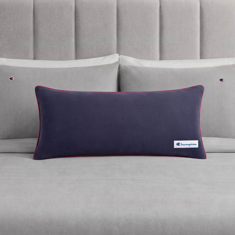 A Champion Faux Wool Body Pillow in navy blue, featuring a small Champion logo, is placed on a neatly made bed with grey sheets and two matching grey pillows, each adorned with a subtle Champion accent.