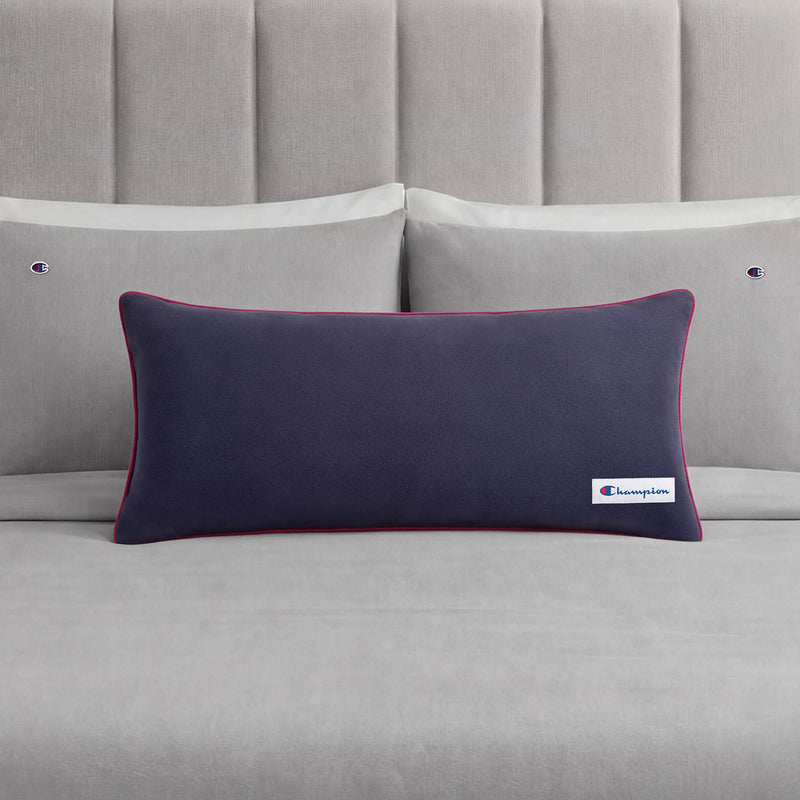 A Champion Faux Wool Body Pillow in navy blue, featuring a small Champion logo, is placed on a neatly made bed with grey sheets and two matching grey pillows, each adorned with a subtle Champion accent.