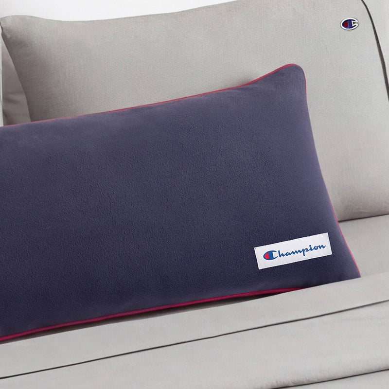 A Champion Faux Wool Body Pillow in navy blue with red trim rests on a light gray couch, and both the pillow and the couch display the Champion logo.