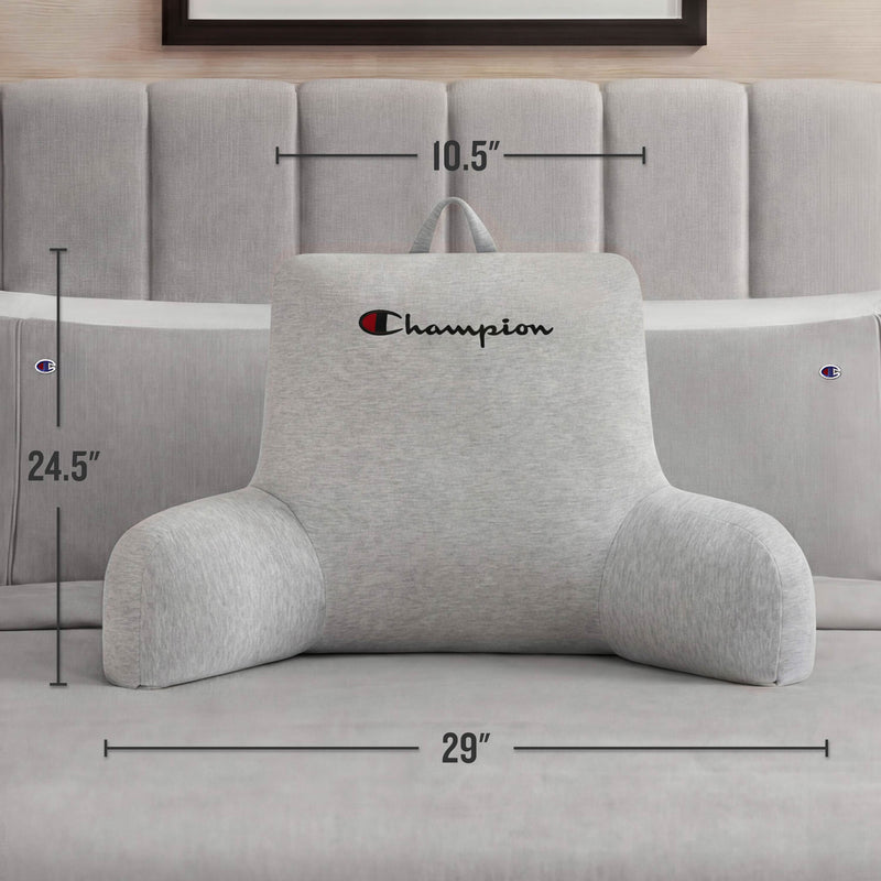 A Champion Jersey Backrest Pillow in gray from Champion rests comfortably on a bed, its arms providing support. The dimensions—24.5 inches tall, 29 inches wide, and 10.5 inches deep—are clearly noted. This cozy pillow is adorned with the iconic Champion logo and placed against a tufted headboard.