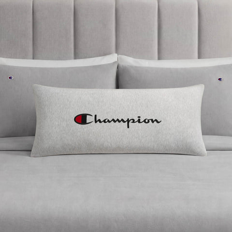 A Champion Jersey Body Pillow in gray, featuring the iconic Champion logo and branding, rests on a coordinating gray bed with two matching pillows and a padded headboard. Made by Champion from soft jersey-knit polyester, this body pillow offers full-length cushioning and comfort for your sleep space.
