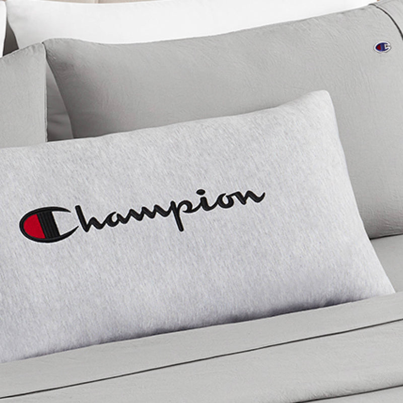 A close-up of a Champion Jersey Body Pillow in gray jersey-knit polyester, showcasing the iconic Champion logo and text, rests on a matching gray bed alongside another pillow featuring the brand’s emblem. This cozy setup, with its full-length cushioning, is perfect for relaxing with your favorite Champion body pillow.
