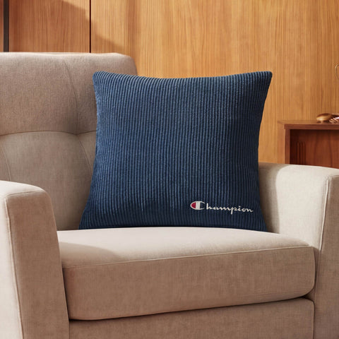 A beige armchair is accented with a Champion 18" x 18" Logo Pillow in blue corduroy, showcasing the iconic Champion logo, set against wooden walls and furniture for a relaxed, casual vibe.