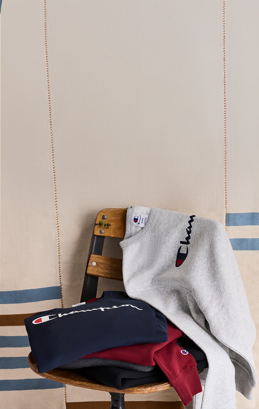 Three sweatshirts in navy, red, and gray on a wooden chair against a striped wall.
