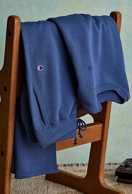 Blue sweatshirt with Champion logo draped over a wooden chair against a light blue wall.