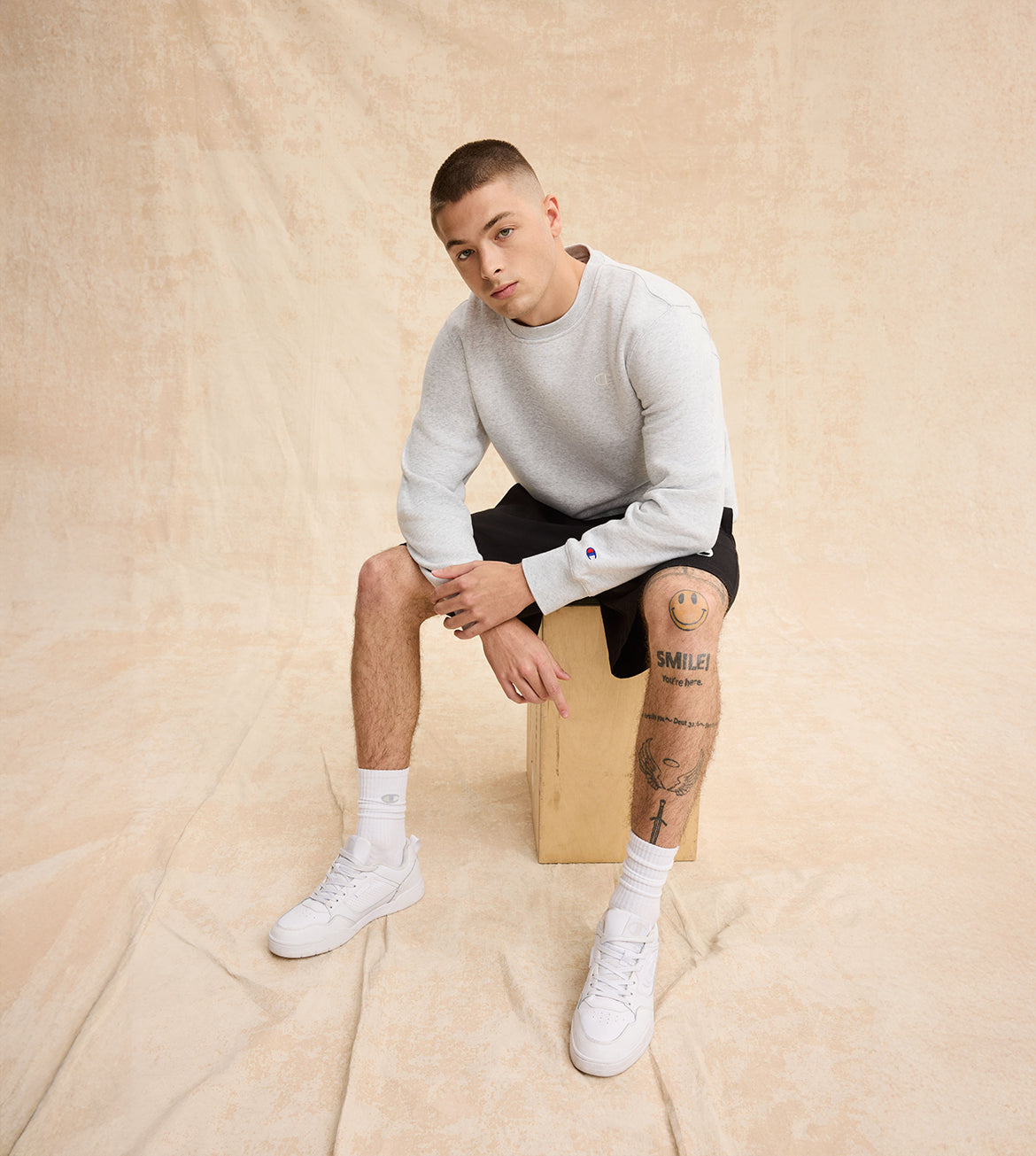 Man sitting on a box against a beige background wearing a white sweater, black shorts, and white sneakers.