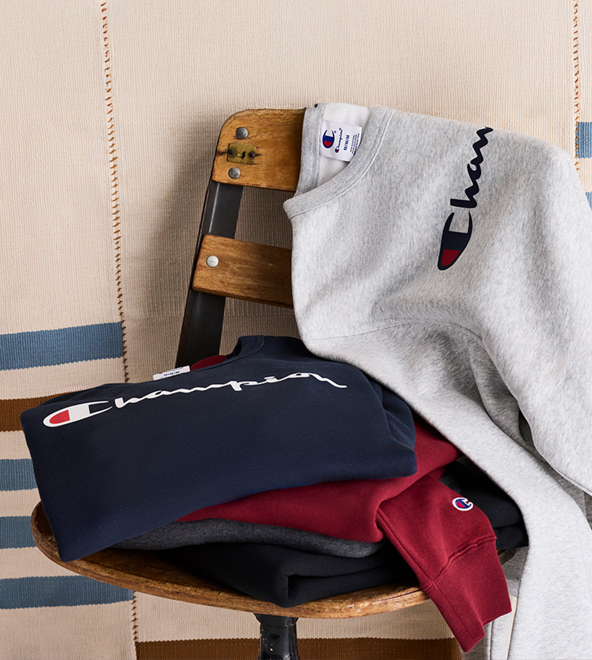 Stack of Champion branded sweatshirts on a wooden chair with a striped rug in the background.