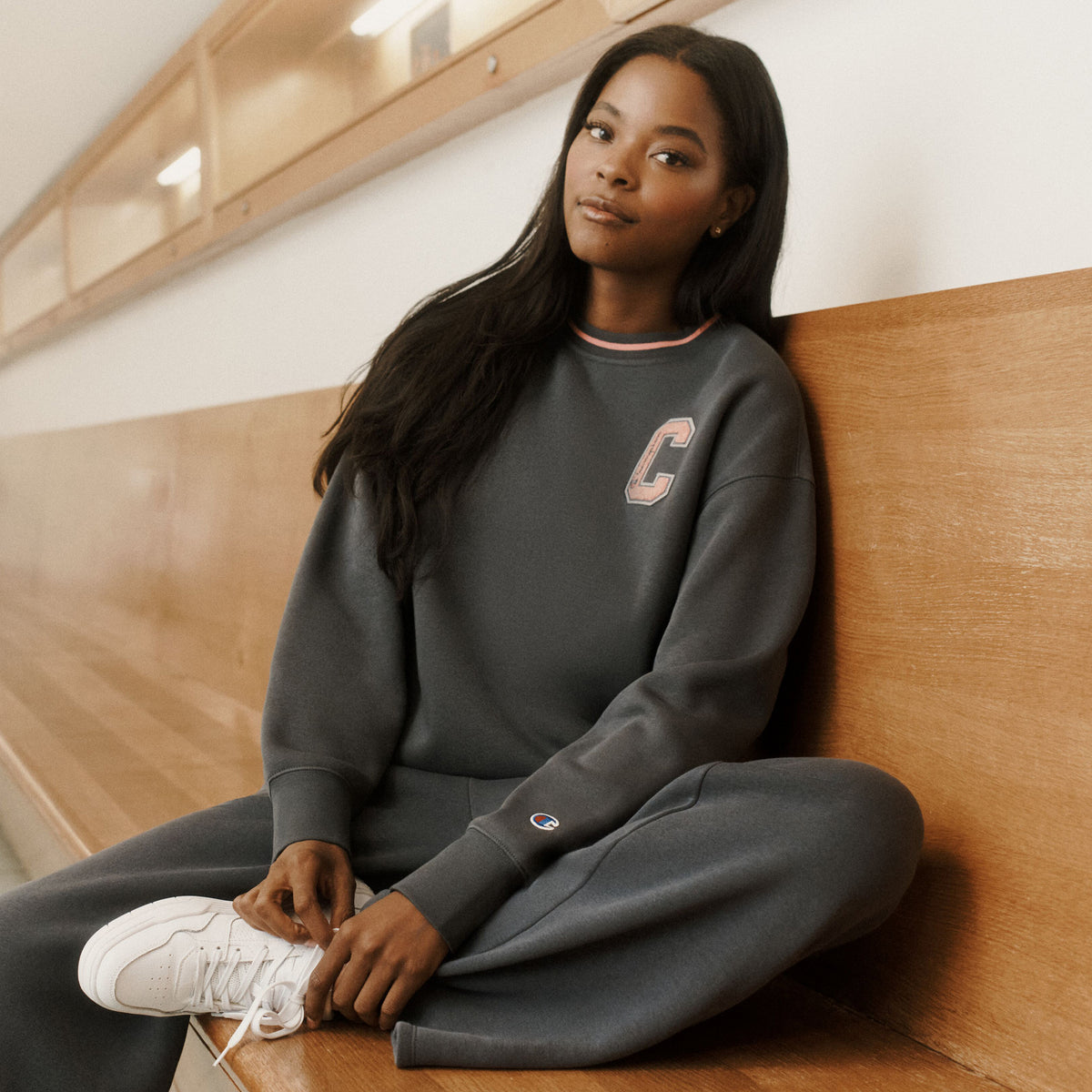 A woman sits on a wooden bench wearing a dark gray Champion sweatshirt and matching sweatpants, with a large pink 'C' on the chest and a small Champion logo on the sleeve cuff. She is also wearing white sneakers.