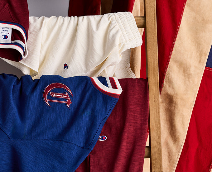 Collection of Champion branded clothing including a blue shirt, red shorts, and white pants on a wooden rack.