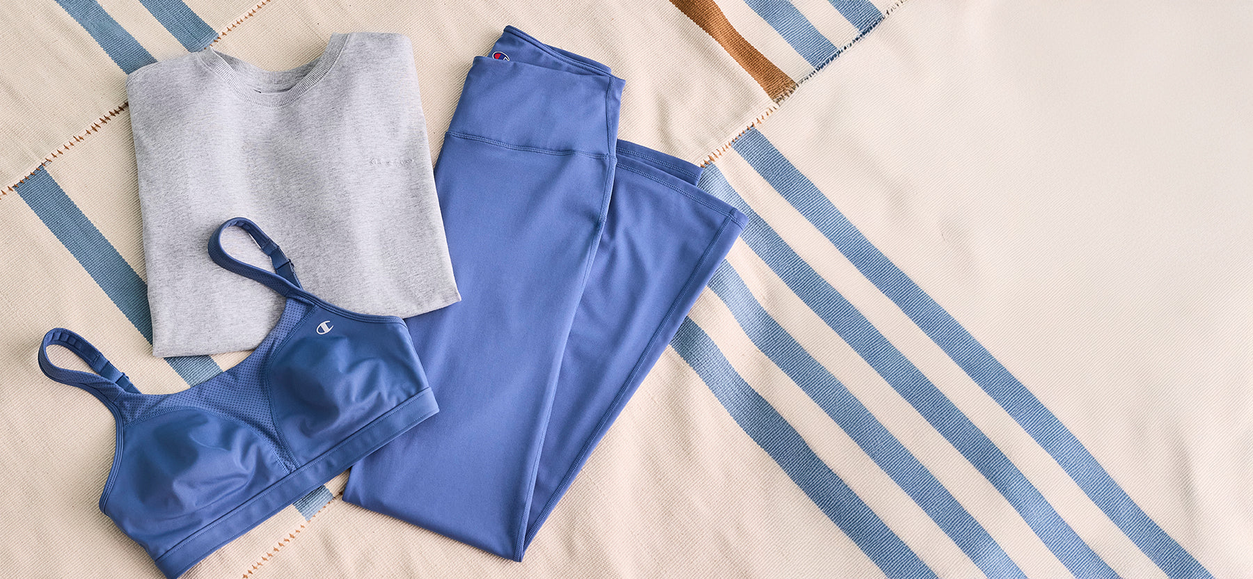Flat lay of a folded gray Champion sweatshirt, blue sports bra, and matching blue leggings arranged on a striped blanket.