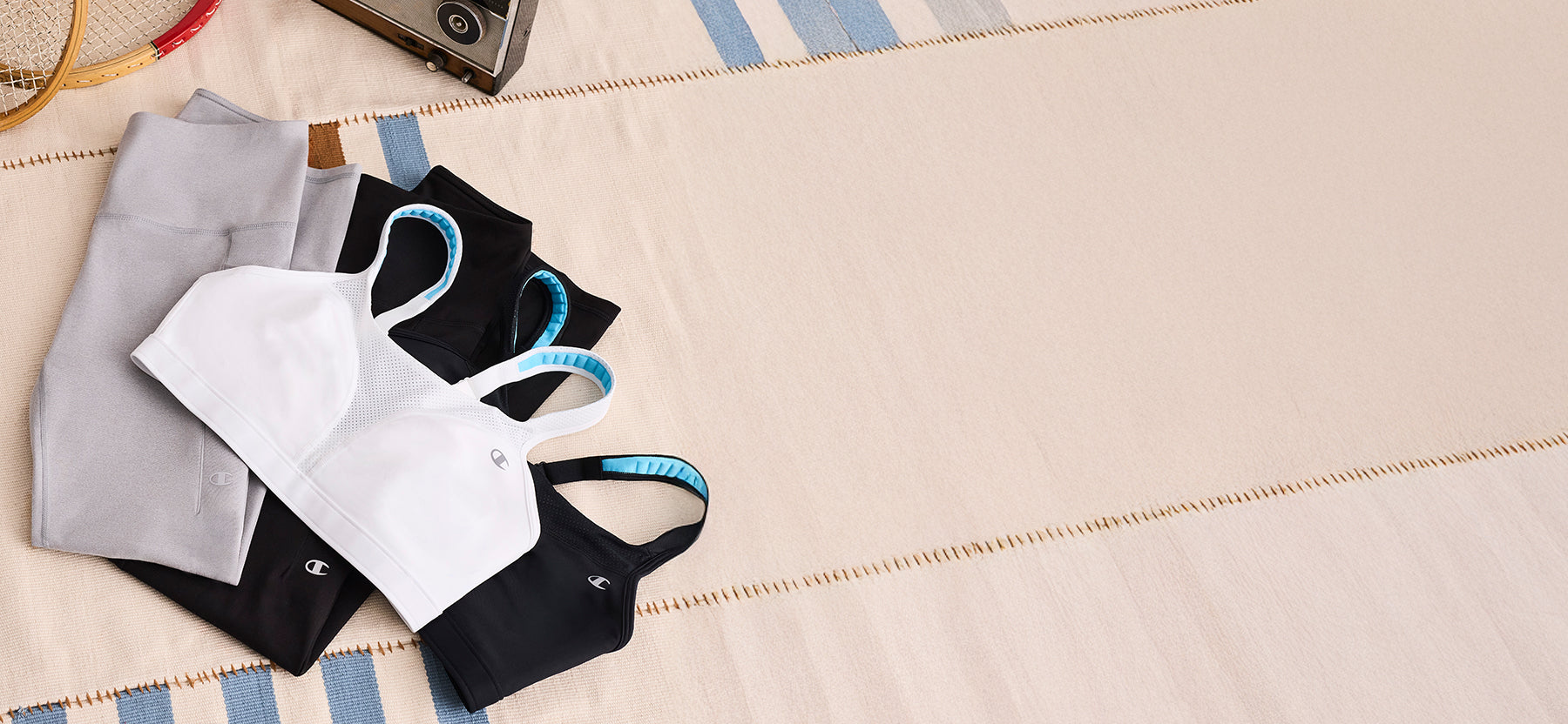Three sports bra cups in white, black, and gray are arranged on a beige fabric surface with a blue-and-white striped mat. A small metallic camera is partially visible in the top corner.