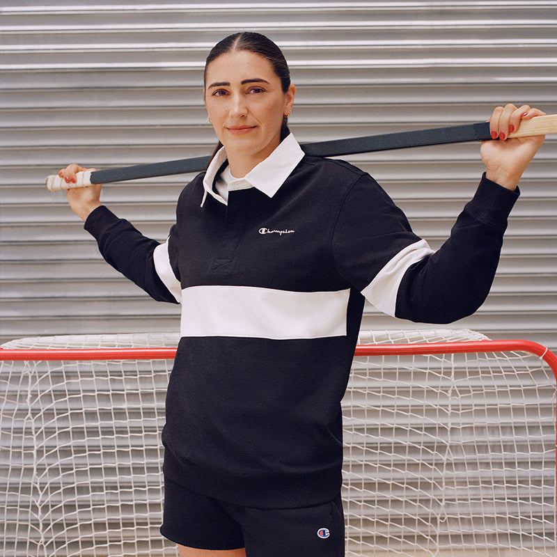 A woman wearing a black and white rugby-style collared top and shorts stands in front of a sports net, holding a hockey stick over her shoulders. The top has a white collar and a wide white horizontal band, with a small visible Champion logo on the chest.
