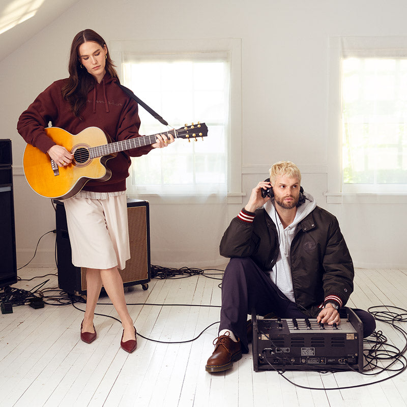 A woman stands holding a yellow acoustic guitar in a bright room with white walls and windows, while a man sits on the floor using a keyboard and phone in front of speakers and cables.