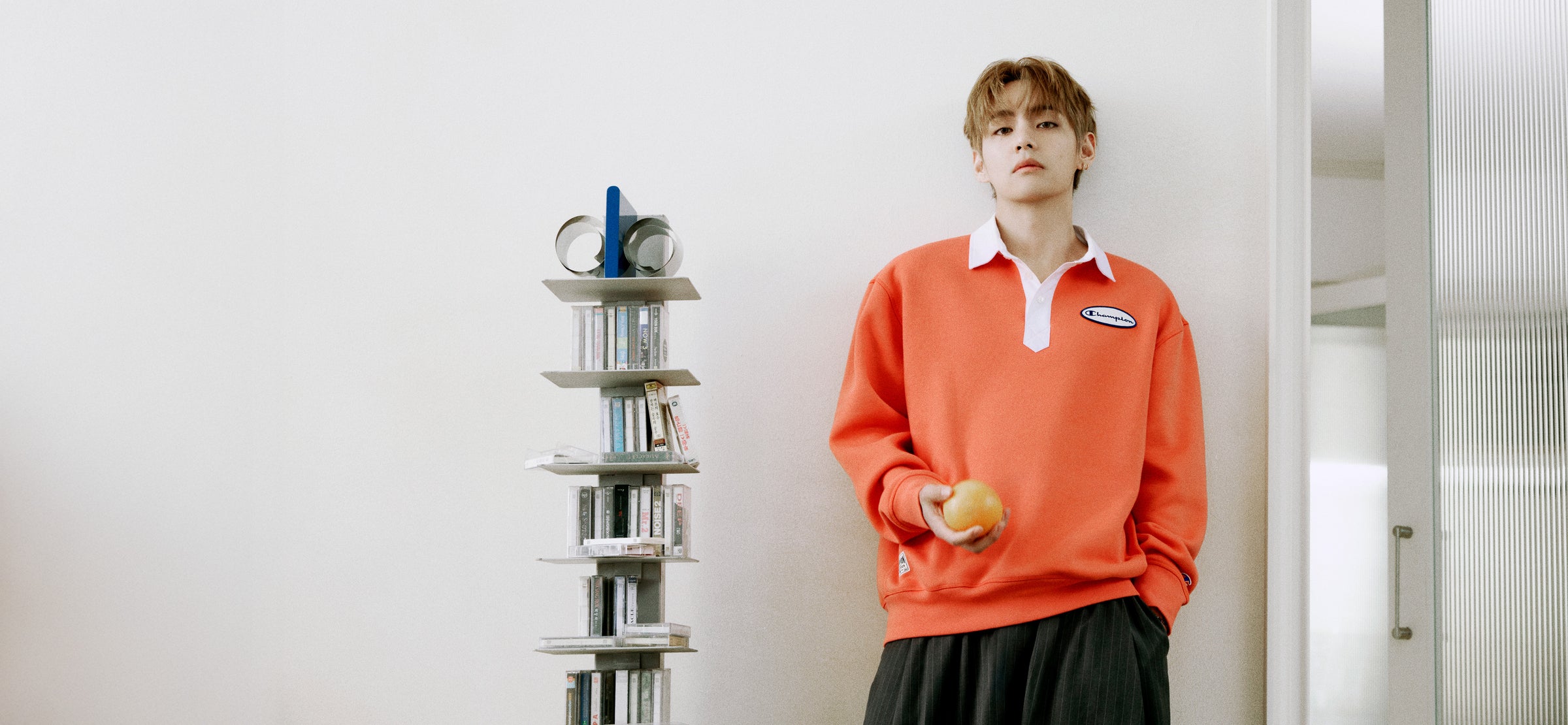 A young man stands indoors against a white wall wearing a bright orange long-sleeve polo shirt with a white collar and a small blue-and-white chest patch, paired with dark pants. A tall narrow bookshelf with books and small objects is beside him.