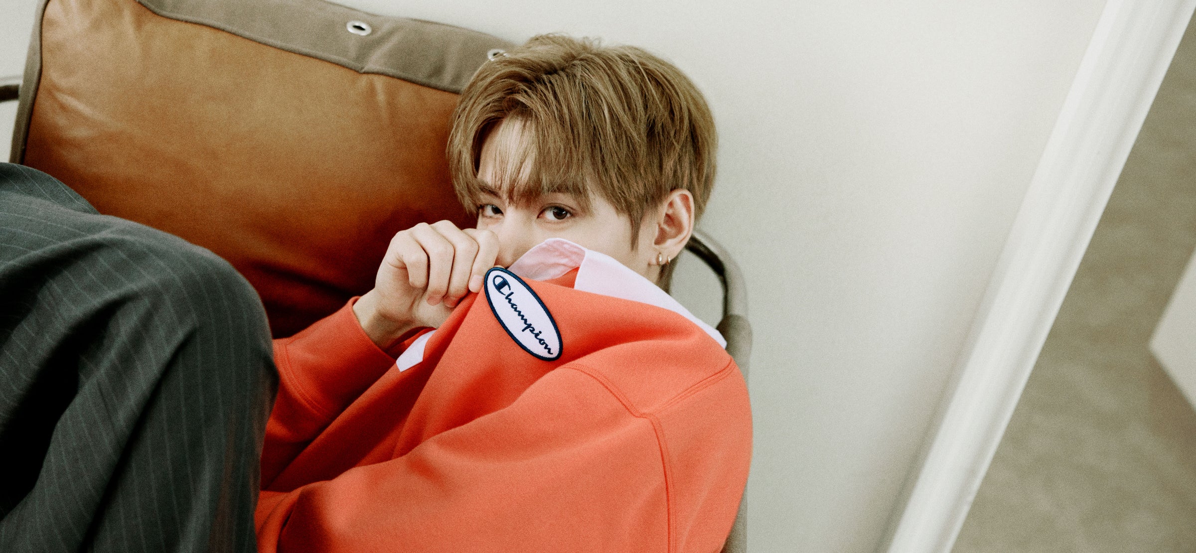 A person wearing a bright orange sweatshirt with a visible oval Champion logo patch on the sleeve, leaning against a brown leather couch in a real indoor setting.