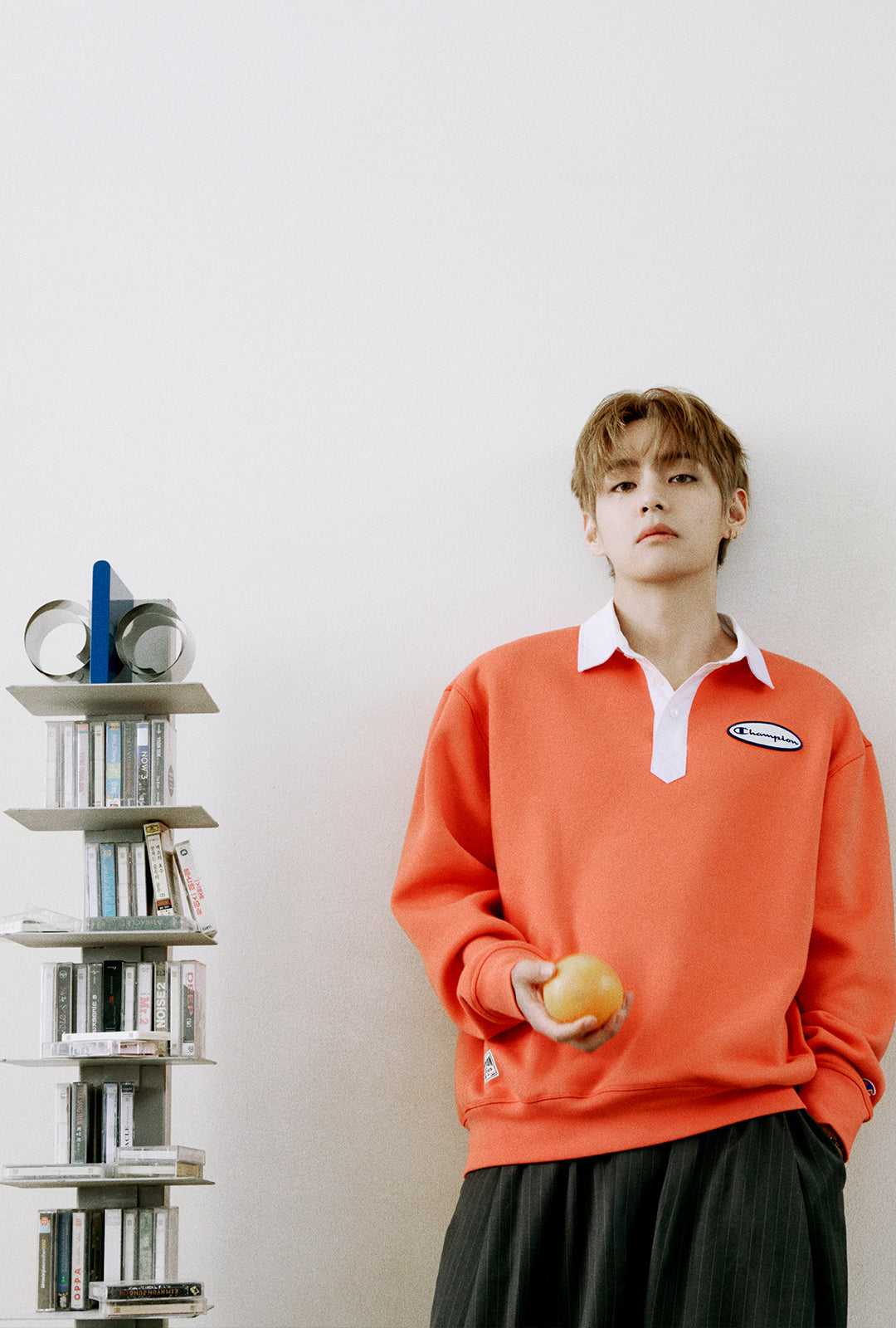 A young man stands against a plain light wall holding a small round fruit, wearing a bright orange long-sleeve polo-style sweater over a white collared shirt and dark pleated skirt. A small bookshelf with books and cups is visible to his left.