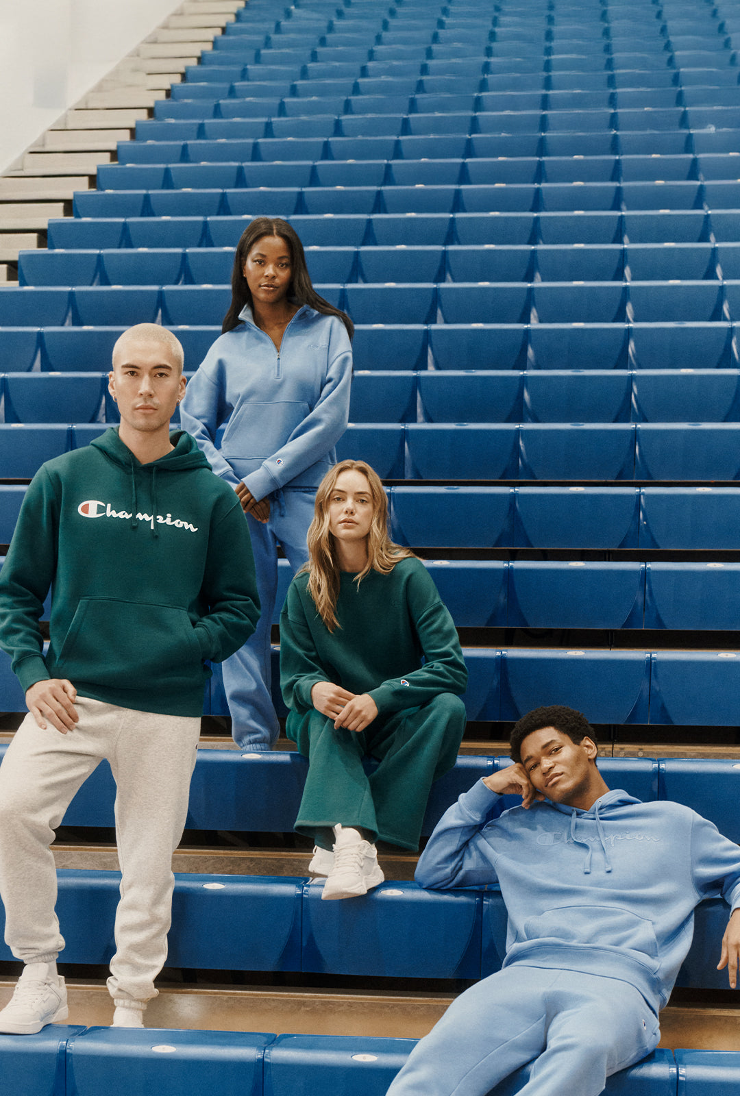 Four models wearing matching Champion sweatshirt and jogger sets in green, blue, and light blue, posed on blue stadium seating.