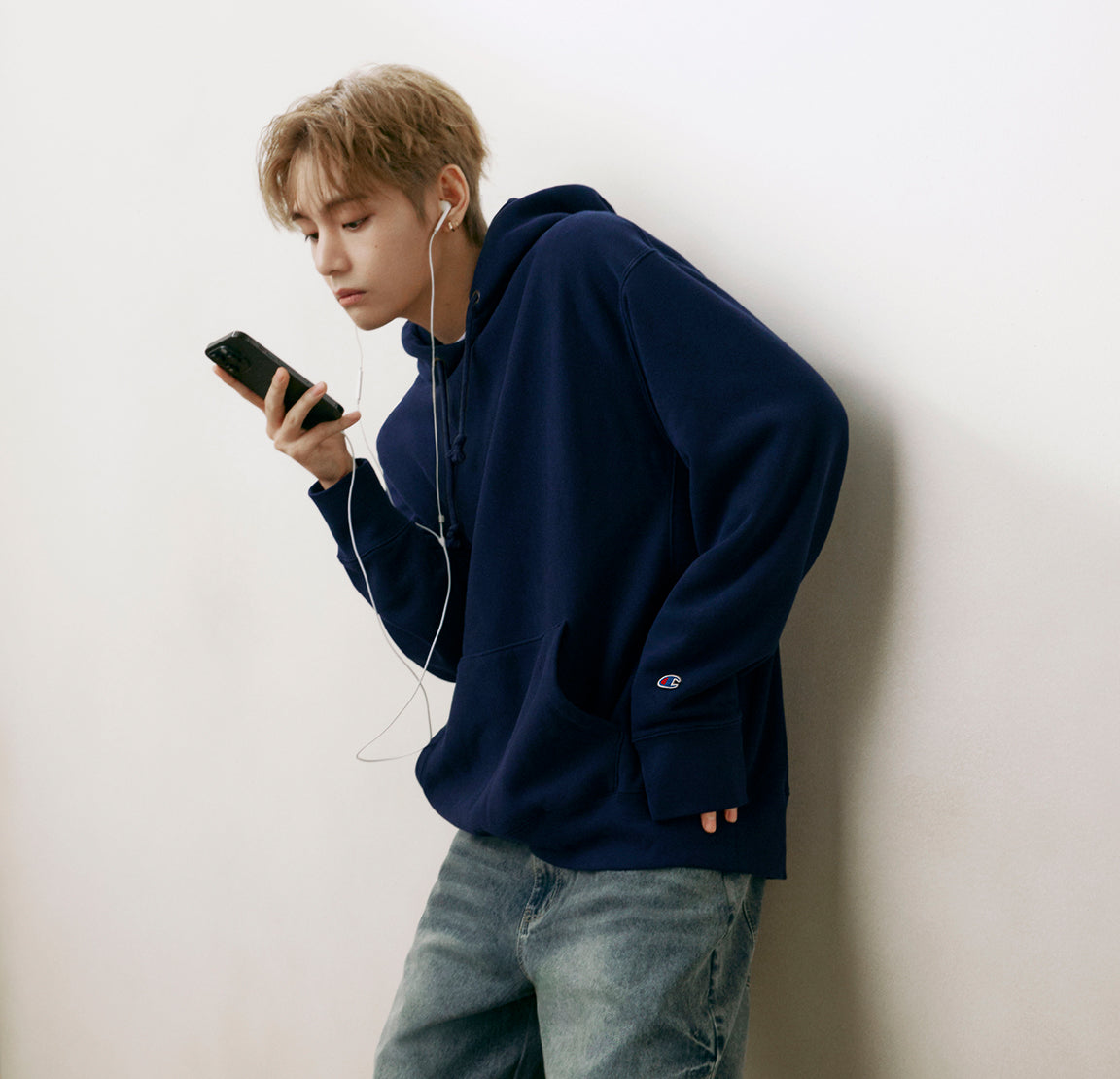 A young man with light brown hair leans against a white wall, wearing a navy blue Champion hoodie and light wash blue jeans, holding a black smartphone and wearing white earphones.