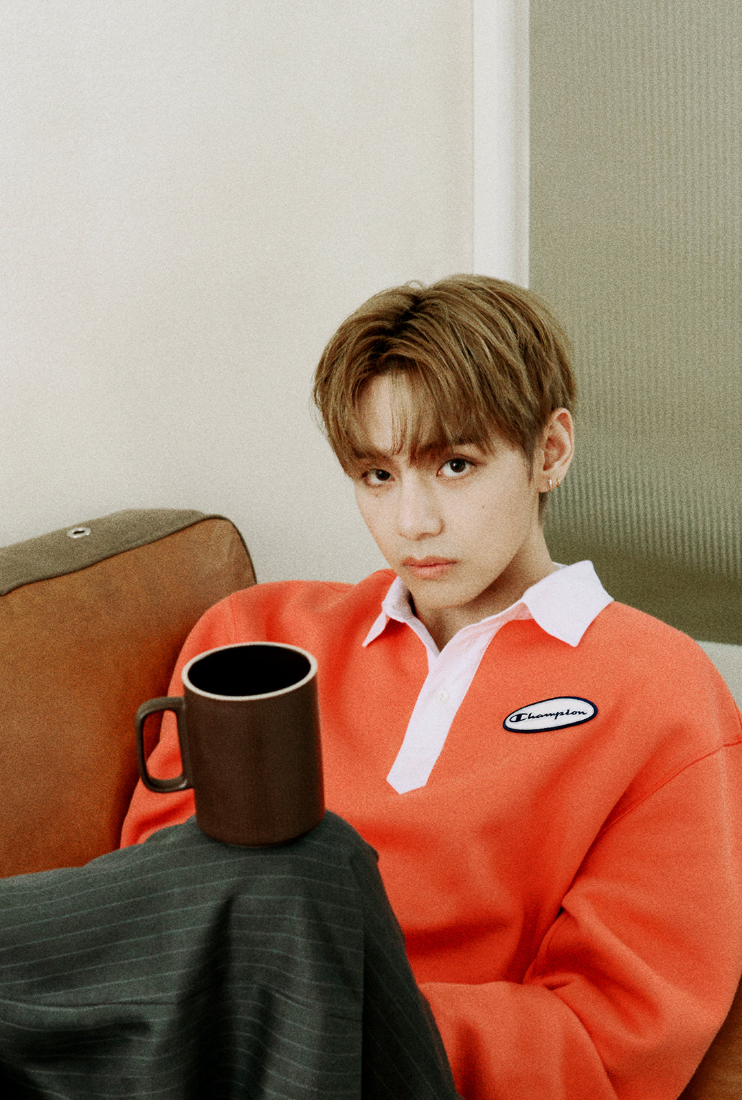 A young man with light brown hair sits on a brown leather couch, wearing a bright orange long-sleeve polo sweater with a white collar and a small oval Champion logo patch on the chest. He is holding a dark brown ceramic mug.