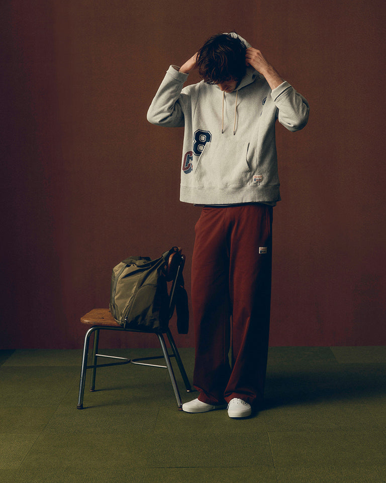 A person stands in a studio setting wearing a light gray hoodie with large navy and blue graphic letters on the front, loose burgundy pants, and white sneakers. A chair with a green bag sits beside them against a dark brown backdrop.