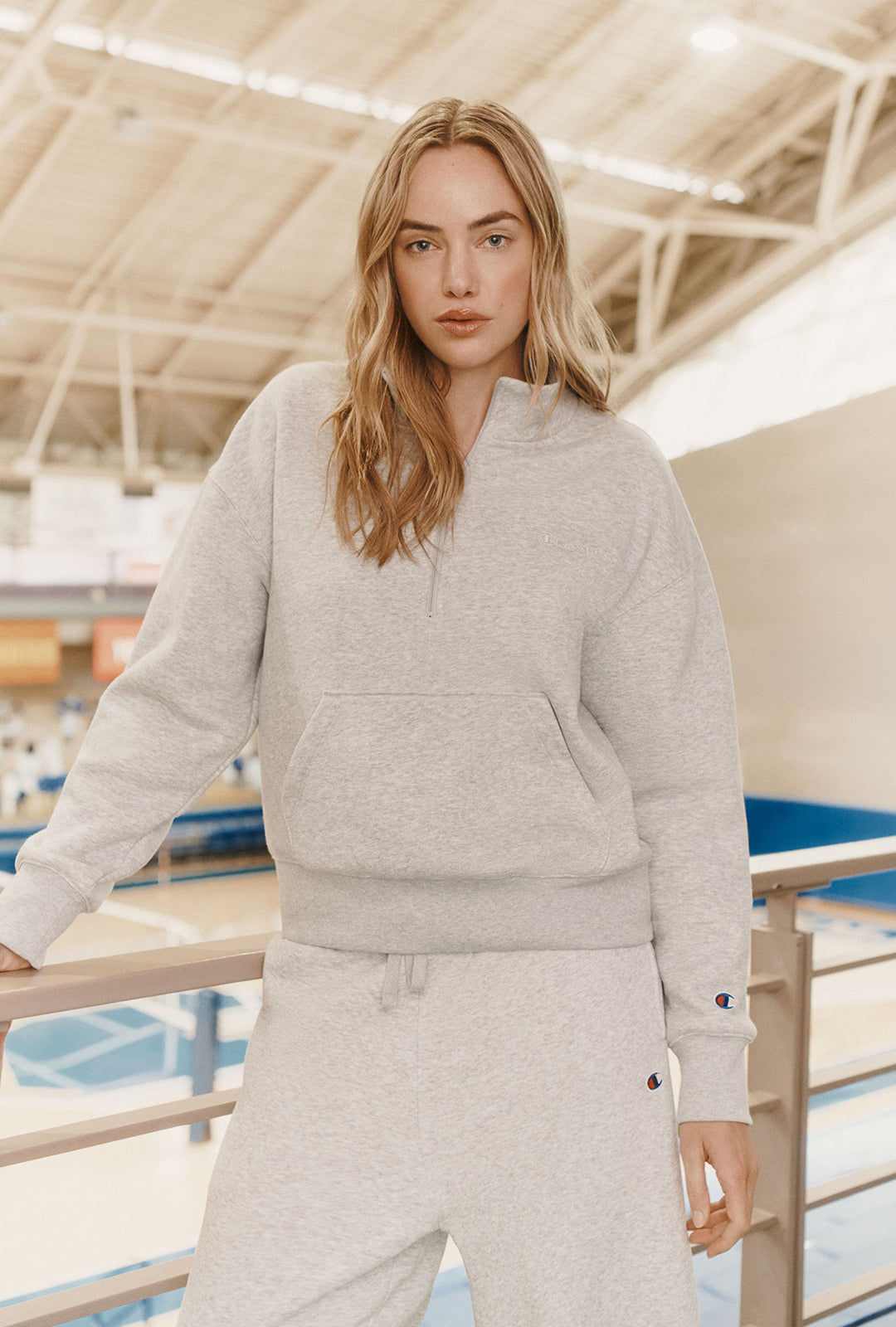 A woman wearing a light heather gray pullover hoodie and matching jogger pants stands indoors near a railing, with a blurred architectural background.