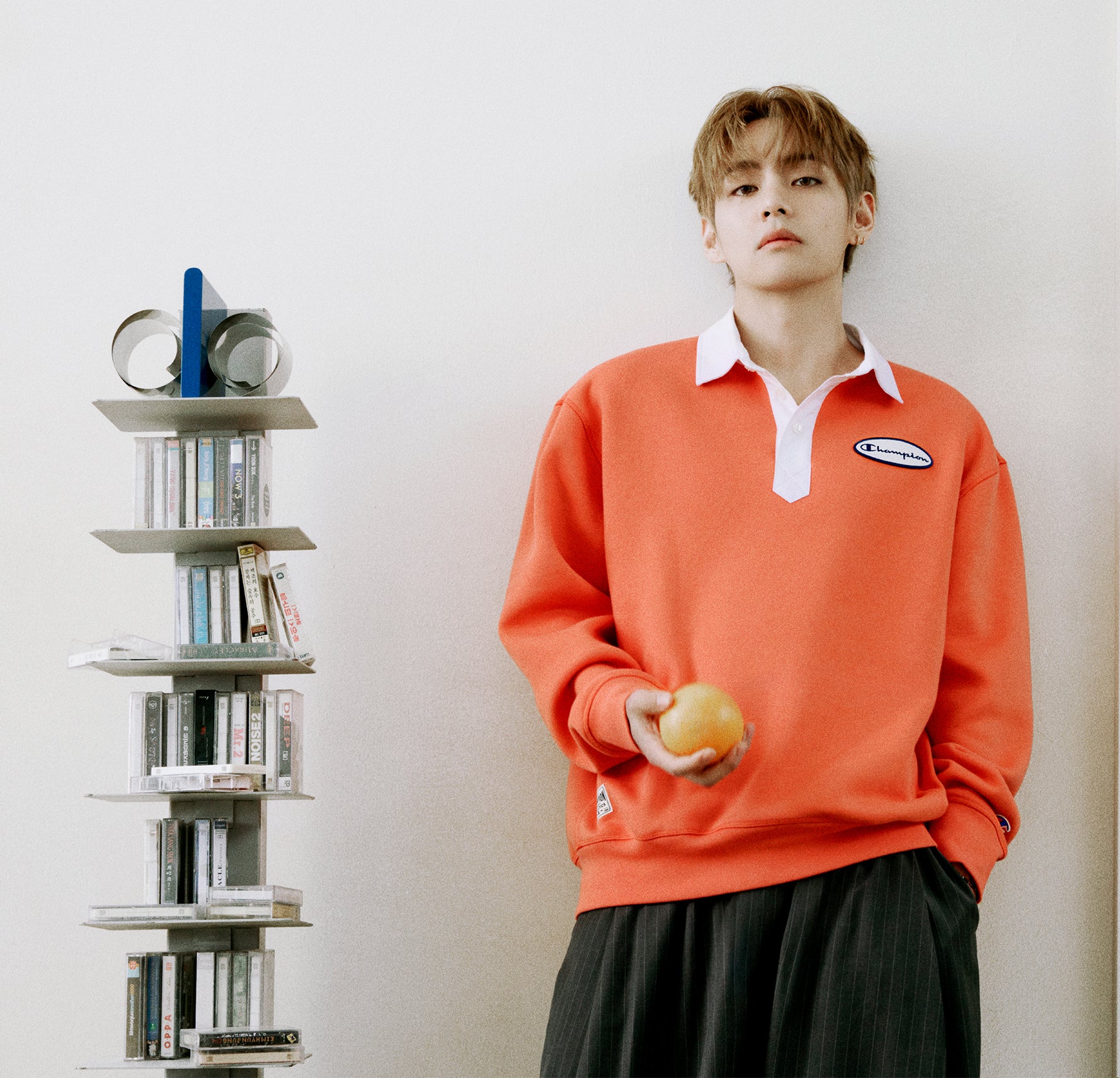 A young man stands against a light wall holding an orange, wearing a bright coral-orange long-sleeve polo sweatshirt over a white collared shirt and dark pleated trousers. To his left is a tall, narrow white bookshelf filled with books and decorative items.