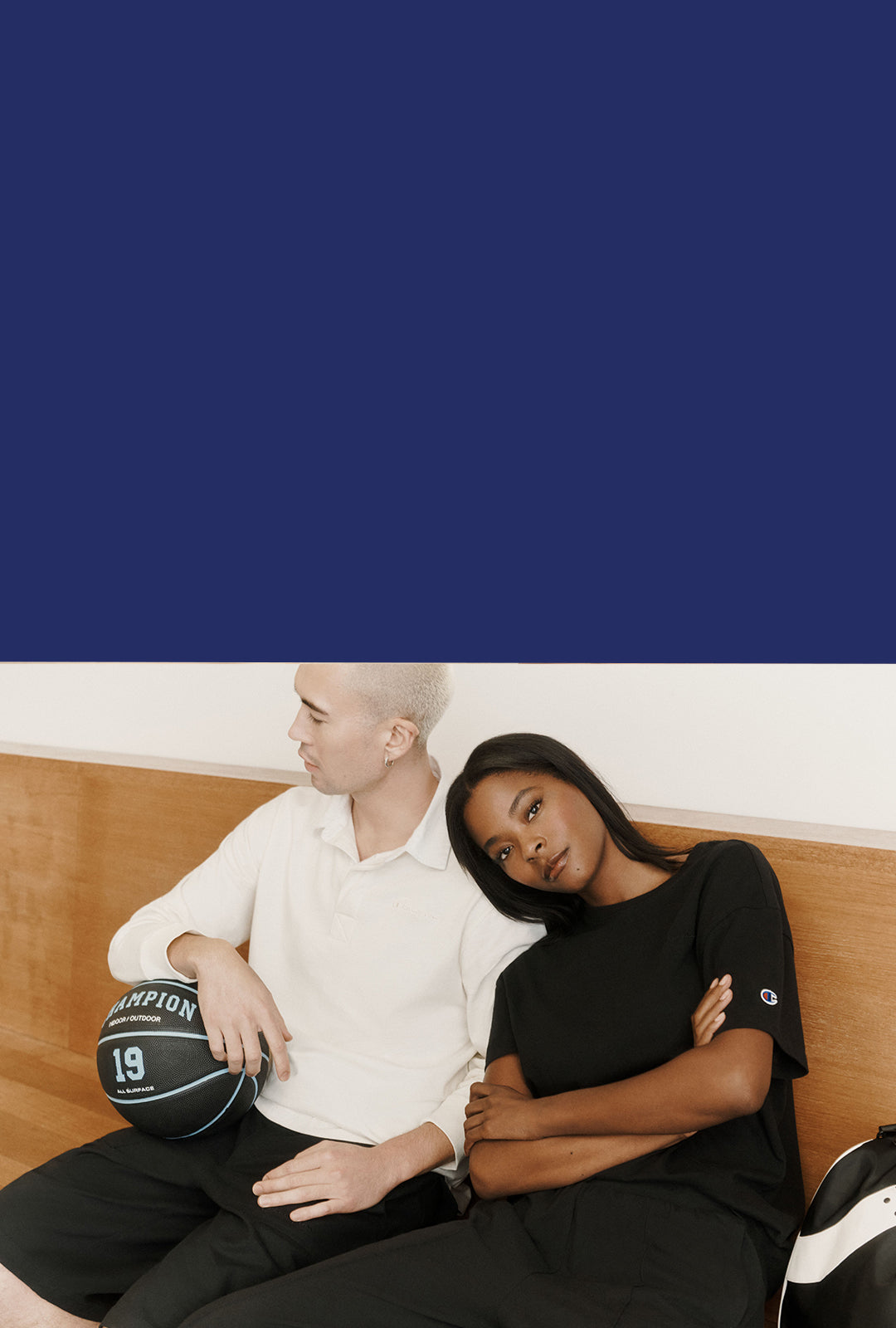 Two models sitting on a wooden bench, one wearing a white polo shirt and holding a black basketball with 'Champion' and the number 19, and the other wearing a black Champion t-shirt and black pants.