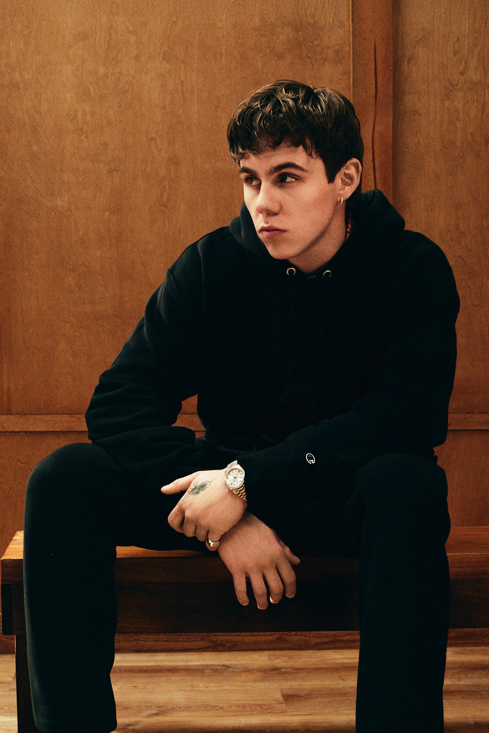 Person wearing a black hoodie and sweatpants sitting on a wooden bench against a wooden wall.
