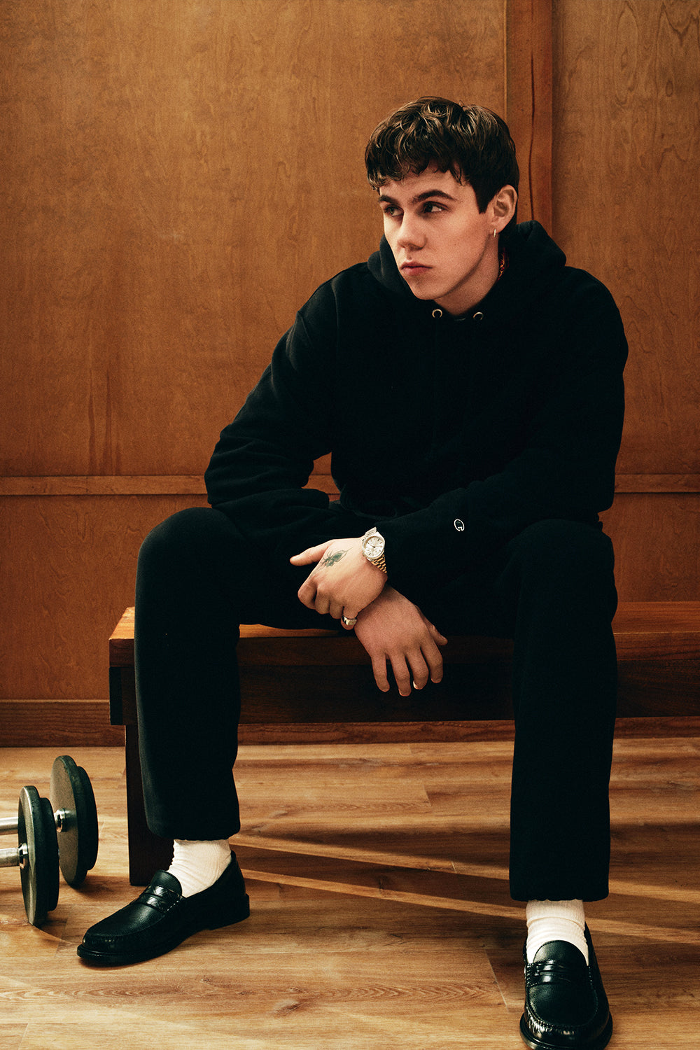 Man in a black outfit sitting on a wooden bench against a brown wall.