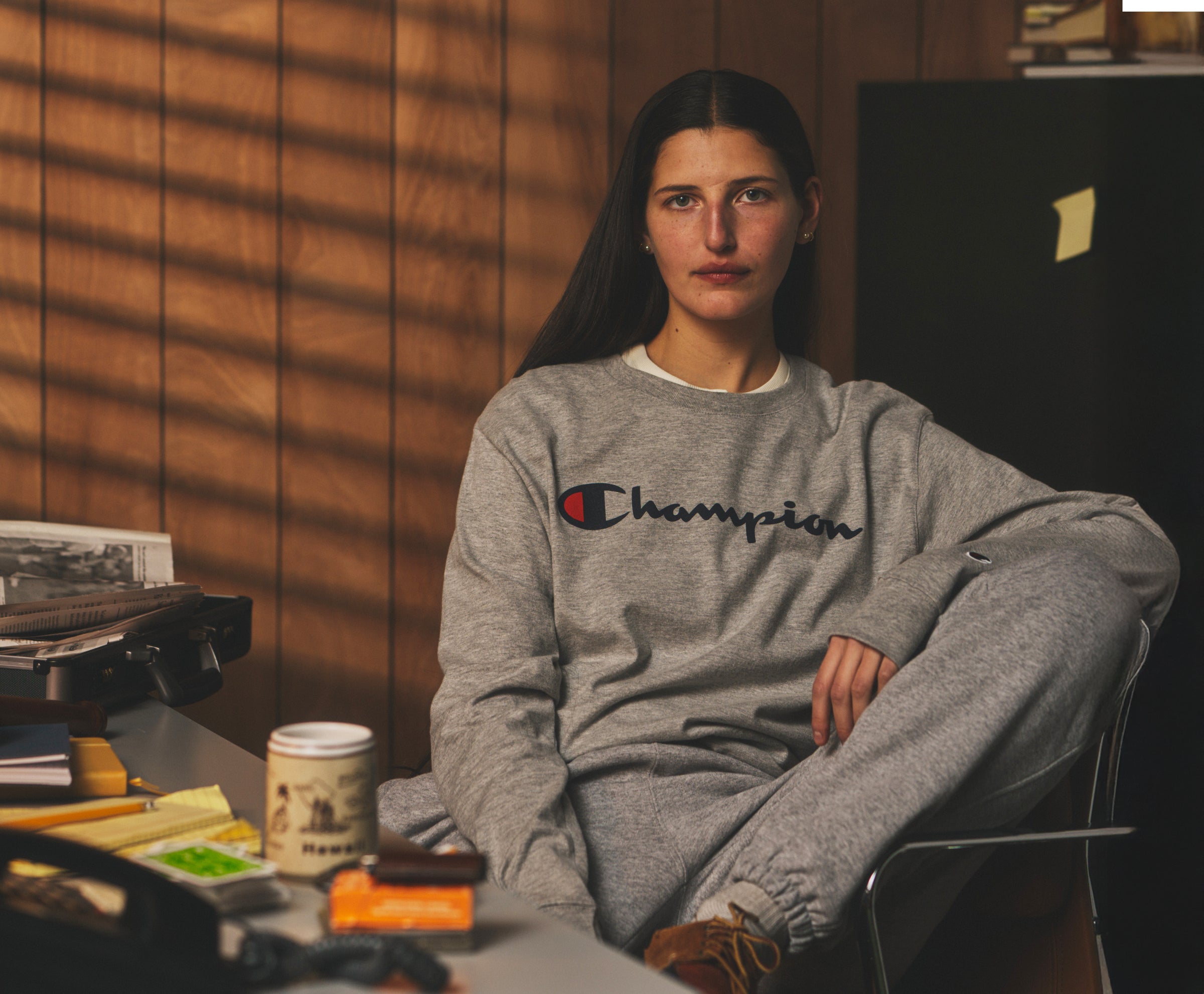 Person wearing a Champion sweatshirt sitting in a room with wooden walls and various items on a table.