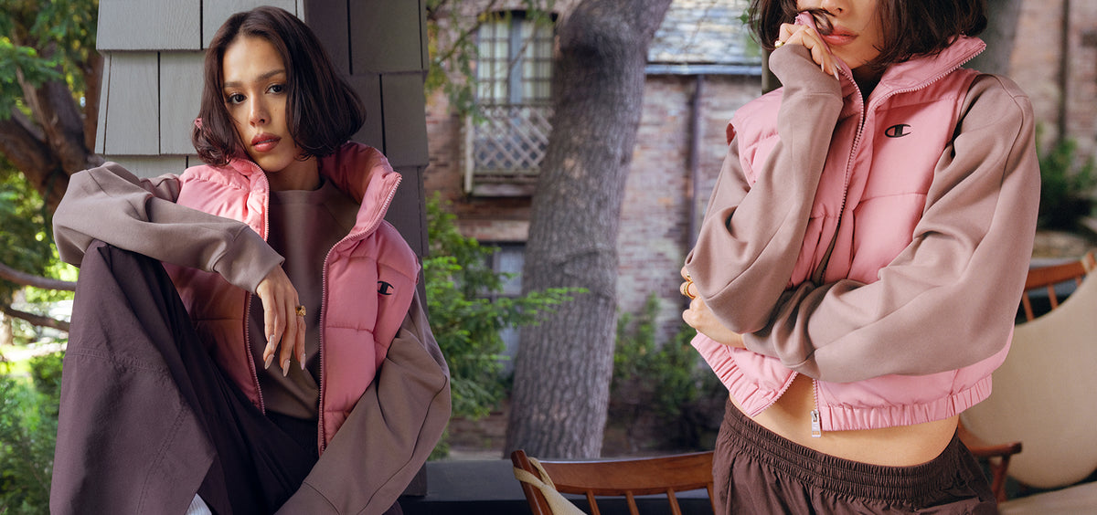 A model is shown in two side-by-side views wearing a pink puffer vest layered over a dusty pink cropped sweatshirt and brown pants, set against an outdoor patio background with trees and a brick building.