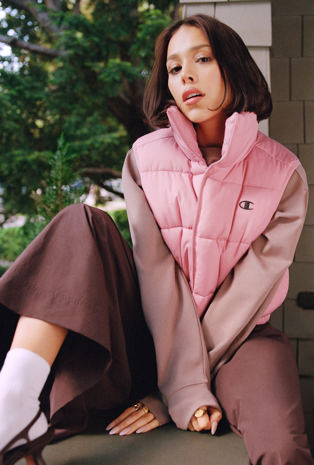 A woman wearing a pink puffer vest with a small embroidered Champion logo, styled with brown wide-leg pants and white socks, sitting outdoors.