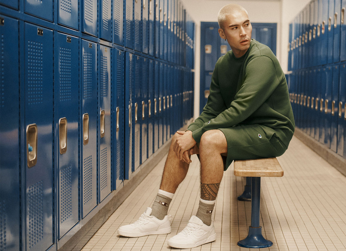A man sits on a wooden bench in a school locker room wearing an olive green crewneck sweatshirt and matching shorts, white sneakers, and gray socks, with blue lockers lining the walls.