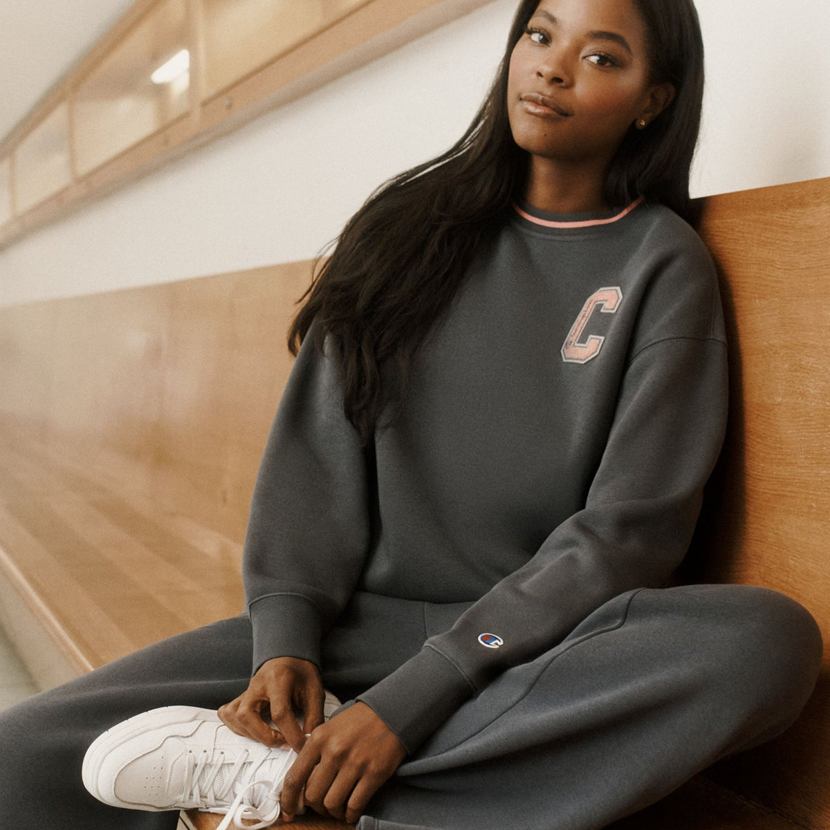 A woman sits on a wooden bench in a bright indoor space, wearing a dark gray Champion sweatshirt and matching sweatpants with white sneakers. The sweatshirt features a light pink embroidered 'C' logo on the left chest and a small Champion logo on the right sleeve cuff.