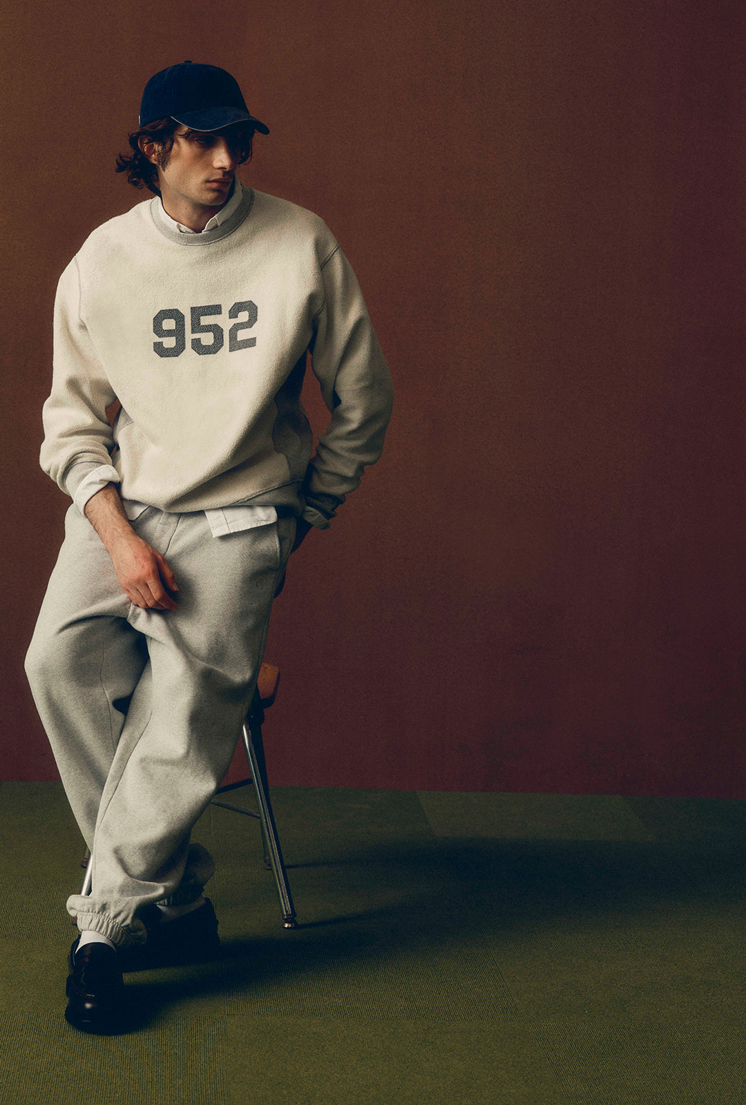 A man wearing a beige sweatshirt with '952' printed on the chest, matching sweatpants, a black cap, and black shoes, leaning against a chair in front of a dark brown wall.