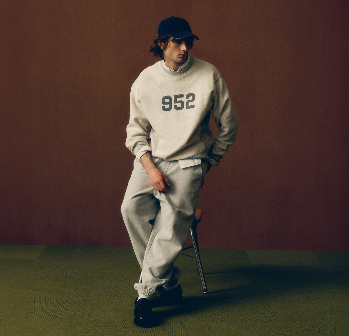 A model wearing a cream sweatshirt with '952' printed on the chest, matching sweatpants, a black cap, and black shoes, leaning against a stool against a solid brown backdrop.