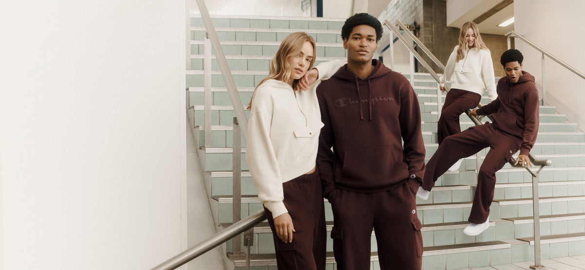Four models wearing matching dark brown tracksuits and a white sweatshirt set, posed on a modern indoor staircase.
