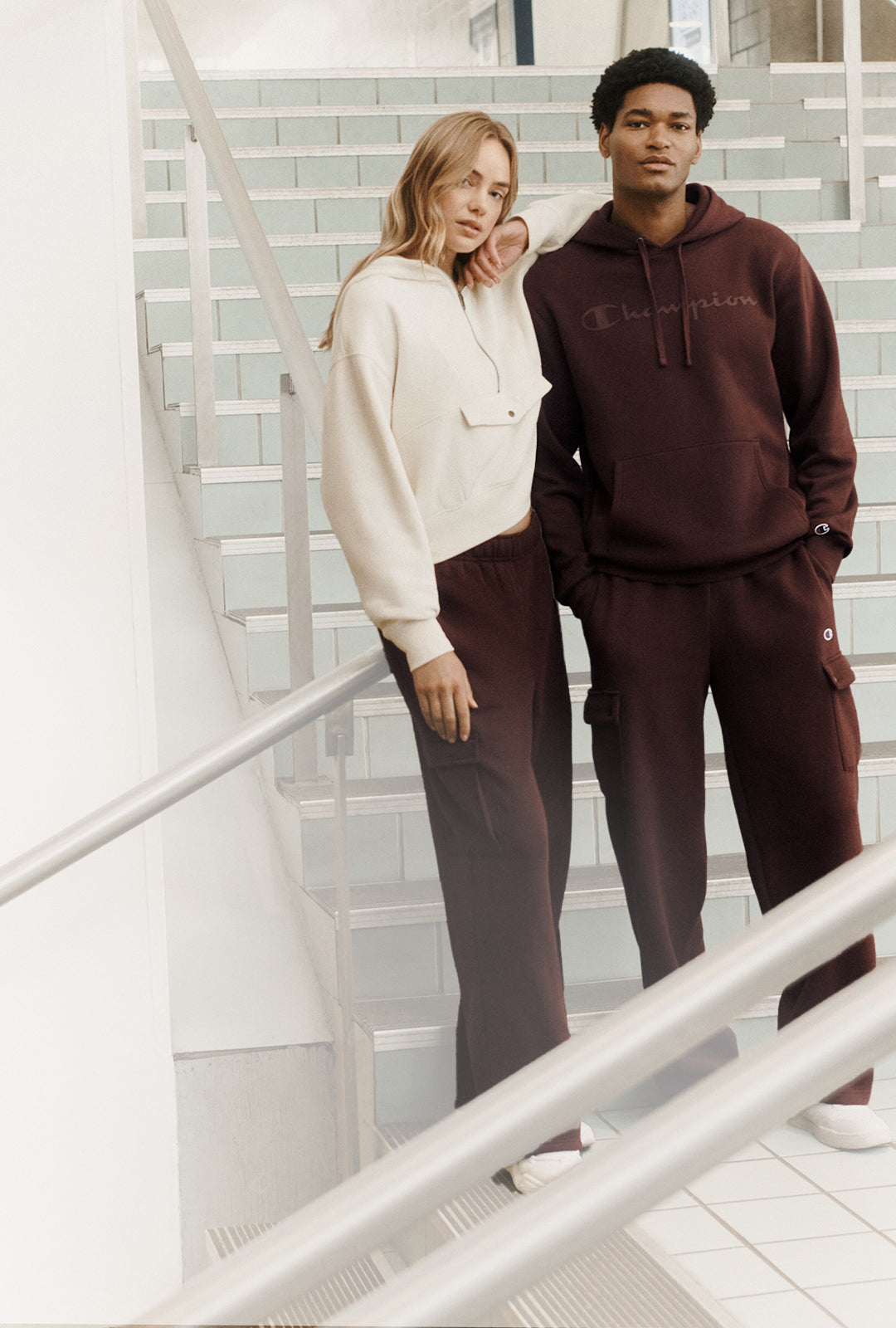A man and a woman modeling matching Champion loungewear sets on an indoor staircase. The woman wears a cream cropped hoodie with dark brown pants, and the man wears a dark brown hoodie with matching pants.