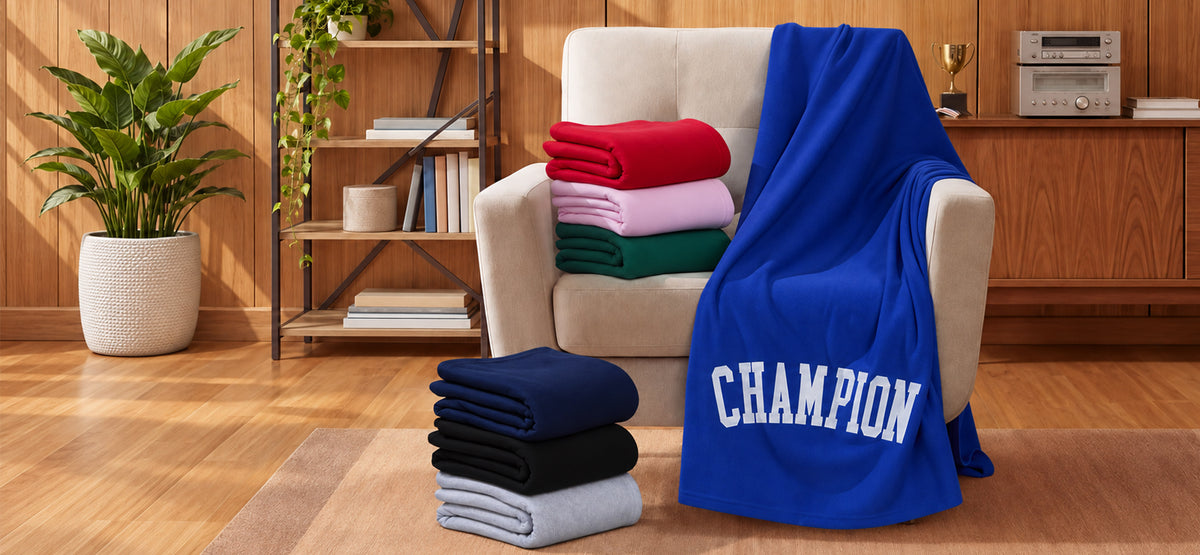 A set of folded blankets in multiple colors, including royal blue, red, pink, green, navy, black, and light gray, displayed on a beige armchair and a wooden floor in a cozy room setting. The royal blue blanket draped over the chair features the word 'CHAMPION' printed in white varsity lettering.
