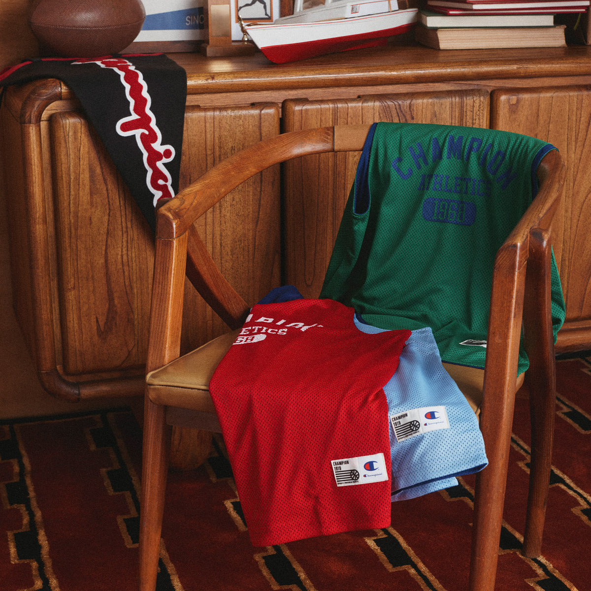 Several Champion athletic jerseys and a scarf are draped over a wooden armchair in a warm indoor room with wood paneling and a patterned rug. Visible products include a red jersey, a green jersey, a light blue jersey, and a black-and-red scarf, all showing the Champion logo.