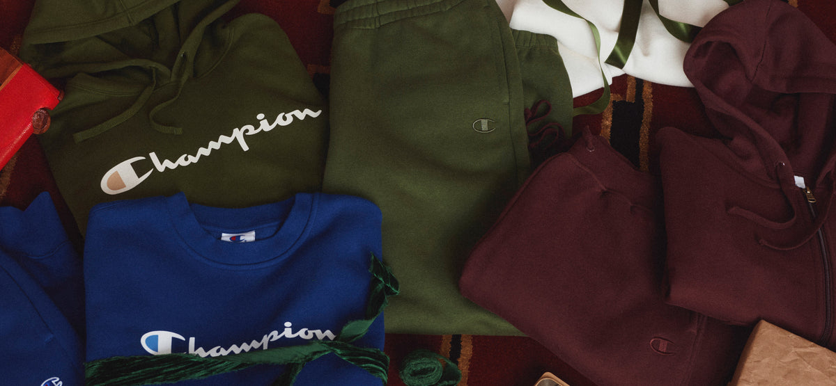 A styled flat-lay of multiple folded Champion apparel items, including an olive green sweatshirt, a royal blue sweatshirt, an olive green pair of sweatpants, and a burgundy zip-up hoodie, arranged on a patterned red and brown rug.